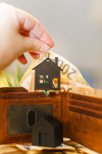 A hand holds a black house-shaped keyring with a green key attached, dangling above an open wooden money box filled with British banknotes. The scene features a close-up of the keyring with the backgr