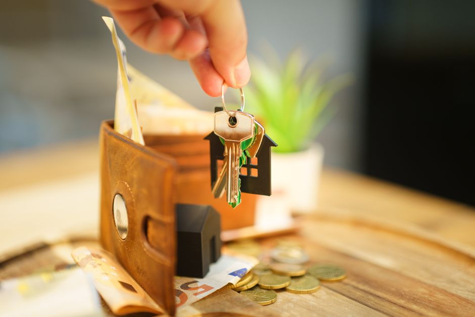 A hand holding a small keyring with three keys and a miniature black house-shaped charm, positioned above a wooden tray containing a cloudy glass vase, some coins, and a partially visible banknote. Th