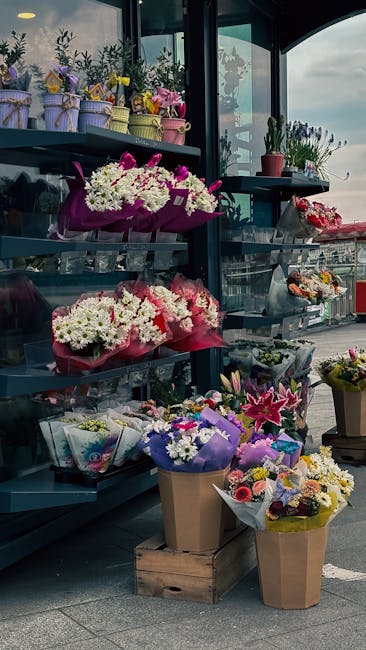 A display of various floral arrangements and bouquets outside a flower shop, featuring vibrant colors and fresh flowers. On the top shelf, small pastel-colored pots with purple, yellow, and pink flowe