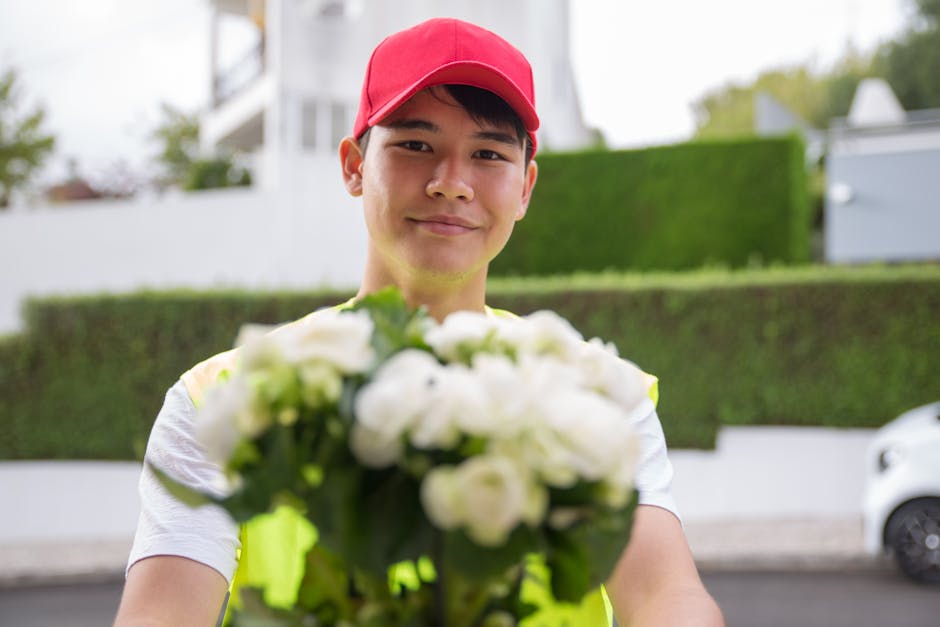 A delivery person wearing a red cap and a high-visibility vest is smiling and holding out a bouquet of fresh white flowers towards the camera. The flowers are arranged in a compact, rounded bouquet wi